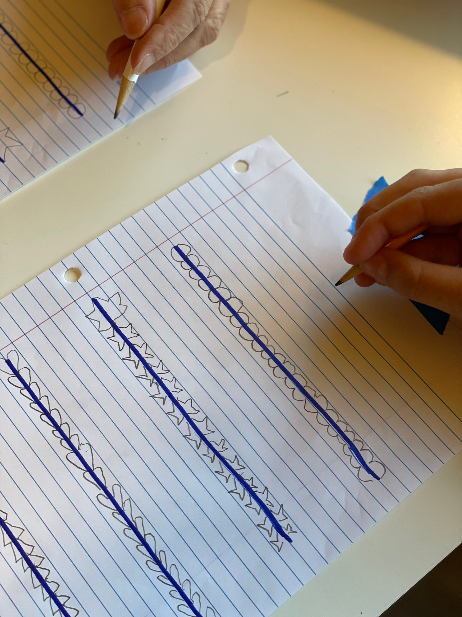 Two people practicing drawing patterns with pencils on lined paper, featuring various shapes like scallops and stars.