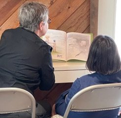 An adult and a child sit together, reading a large open picture book in a room with wood paneling.